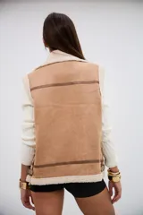 Chaleco de gamuza sintética color camel con interior y bordes de corderito sintético color crema. Presenta cierre frontal metálico, detalles de tiras con hebillas en el cuello y en la parte inferior, y costuras reforzadas con ribetes de cuero sintético.