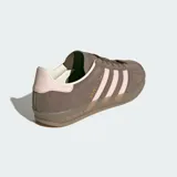 Championes Adidas Gazelle Indoor de diseño clásico, confeccionados en gamuza color marrón con las icónicas tres tiras laterales y refuerzo en el talón en color rosado. Presentan suela de goma translúcida y cierre de cordones.
