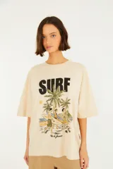 Remera extragrande de algodón color beige, de manga corta y corte holgado. Cuenta con un estampado frontal que dice 'SURF' junto a una ilustración de playa con palmeras, surfistas y la inscripción 'Rio de Janeiro'.