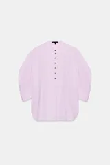 Camisa confeccionada en hilatura de algodón de color rosado claro, con cuello mao y cierre frontal parcial con botones a contraste. Presenta mangas por debajo del codo abullonadas y bajo asimétrico con aberturas laterales.