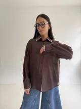 Camisa oversize de color marrón con cuello camisero y puños con detalle de bordado festoneado en color blanco. Presenta cierre frontal de botones y mangas largas.