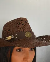 Sombrero de paja trenzada color marrón con forma de sombrero cowboy. Tiene una cinta de cuero sintético con aplique metálico en la base de la copa.