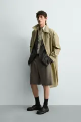 Trench de corte relajado confeccionado en tejido técnico italiano, color beige. Presenta cuello camisero, manga larga, bolsillos de vivo en la cadera y cierre frontal de botonadura simple con botones oscuros.