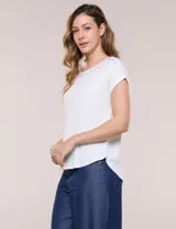 Remera blanca de punto acanalado con escote redondo adornado con tachas metálicas en el cuello y hombros, mangas cortas japonesas y ruedo curvo.
