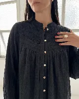 Camisa negra de corte amplio y fluido, con cuello mao, mangas largas y cierre frontal de botones. Presenta un canesú con detalles bordados y calados en la parte superior delantera.