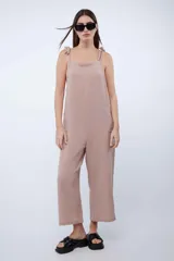 Mono largo de algodón color beige, de corte holgado, con escote recto y tirantes ajustables mediante lazos en los hombros. Cuenta con bolsillos laterales y perneras amplias tipo culotte.