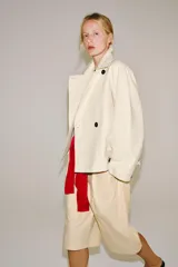 Trench corto color marfil de algodón, con cuello camisero, solapa cortaventos, mangas largas amplias con puños abotonados y cierre cruzado con un botón visible.