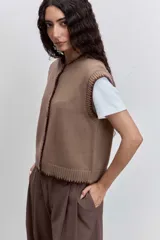 Chaleco de punto color marrón con cierre frontal de botones y detalle de costuras en contraste en los bordes. Presenta un diseño de cuello redondo y corte recto.