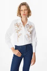 Camisa blanca de corte clásico con cuello camisero y cierre frontal de botones. Presenta un bordado lineal en color marrón oscuro en ambos lados del frente, representando motivos florales.