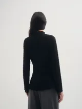 Blusa negra de crepe texturado, con cuello alto, mangas largas acampanadas y corte asimétrico con abertura lateral.