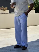 Pantalón de corte ancho y holgado, con diseño de rayas verticales finas en color azul y blanco. Presenta cintura elástica con cordón ajustable y un pequeño bordado en color rojo en la parte superior.