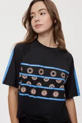 Remera blanca de manga corta, con cuello redondo, calce relajado y estampado frontal con motivos gráficos de soles y símbolos marinos en tonos rosados y bordo, además de líneas horizontales.