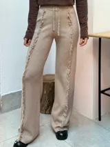 Pantalón de punto color beige, de corte recto y holgado, con cintura elástica y cordón ajustable. Presenta detalles de costuras con flecos verticales a lo largo de las piernas.