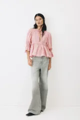Blusa rosada de algodón con ribetes de encaje, cuello redondo con abertura en V y tiras para anudar, mangas largas abullonadas con puños elásticos y terminación peplum con vuelo.