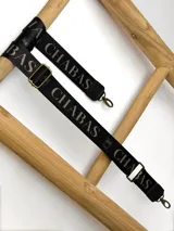 Correa ajustable para cartera confeccionada en cinta tejida con diseño de rayas en negro, blanco y beige, con punteras de cuero sintético negro y herrajes metálicos.