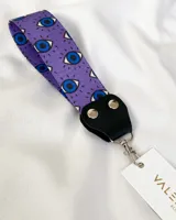 Llavero de tela color lila con estampado de ojos azules y negros. Tiene un conector de cuero sintético negro con remaches plateados y un gancho metálico plateado.
