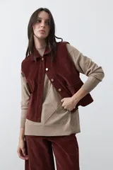 Chaleco de pana color marrón oscuro, con cuello camisero, cierre frontal de botones metálicos dorados y dos bolsillos frontales con detalle de bordado geométrico.