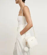 Cartera bandolera Satinato pequeña de color blanco, de diseño estructurado con textura floater, cierre de cremallera y detalles metálicos dorados. Incluye una correa larga ajustable y un pompón decorativo colgante.