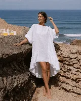 Vestido corto de lino color blanco, con mangas cortas amplias, escote redondo y diseño asimétrico más corto en la parte delantera y con volados en el ruedo.