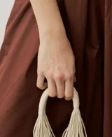 Cartera de mano de macramé color crudo con apliques de caracoles y flecos colgantes en la parte inferior. Cuenta con asas rígidas integradas en el mismo tejido.