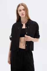 Camisa negra de viscosa, holgada, con mangas cortas abotonadas, cuello camisero, detalles de cintas decorativas verticales y cierre frontal con botones.