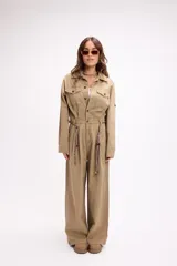 Mono de estilo utilitario color beige, con cierre frontal de botones, bolsillos en el pecho y cintura entallada con detalle de cadenas decorativas. Presenta mangas largas y piernas de corte recto y holgado.