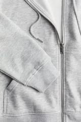 Campera con capucha y cierre frontal, color gris melange, confeccionada en felpa con interior cepillado suave. Presenta hombros caídos, bolsillos canguro y puños y bajo en punto acanalado.