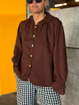 Camisa de lino color marrón con cuello redondo decorado con volados, cierre frontal de botones y mangas largas con puños ajustados.