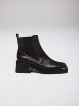 Botas tipo chelsea de cuero color marrón oscuro con acabado brillante. Presentan punta cuadrada con detalle de puntera metálica, elásticos laterales y taco cuadrado bajo.