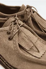 Mocasín de cuero gamuza en color tostado, con cordones, flecos frontales y suela gruesa con costuras visibles.