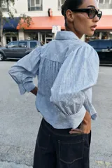 Camisa de cuello solapa y manga larga con puños abotonados. Presenta un diseño de rayas verticales azules y blancas con un estampado floral lineal superpuesto. Cierre frontal con botones.