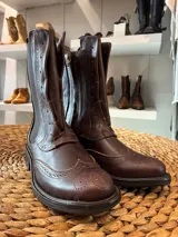 Bota tipo borsego de cuero color chocolate, con diseño estilo brogue y perforaciones decorativas. Cuenta con elástico lateral, cierre interno y suela de goma con taco bajo.