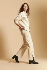 Pantalón de corte recto y tiro medio, color beige, con detalle de tachas metálicas a lo largo de las costuras laterales y tajos en el ruedo.