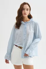 Camisa celeste de algodón con diseño broderie, cuello a la base con volados, botonadura frontal con detalle de volado lateral y mangas largas con puños elastizados y con volados.
