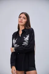 Camisa negra de manga larga con cuello clásico y botones frontales. Presenta dos grandes apliques bordados con lentejuelas y cuentas en forma de flores y hojas en la parte delantera.