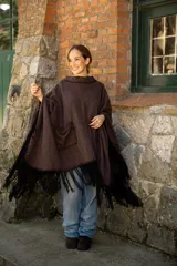 Poncho de tejido suave color marrón chocolate, con diseño amplio y relajado. Presenta detalles artesanales de costura en los bordes y en el bolsillo frontal, además de flecos largos en color negro en todo el ruedo.