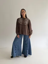 Camisa color marrón chocolate con bordado artesanal de flores y hojas en hilo crudo. Presenta cuello con volado, mangas largas con puños abotonados y cierre frontal con botones.