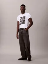 Remera blanca de algodón con cuello redondo y manga corta, presentando un estampado gráfico apilado en negro con el texto "CALVIN KLEIN JEANS".