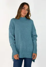 Sweater de punto color azul petróleo, con cuello alto, mangas largas y puños con detalle de tejido trenzado. Presenta un diseño holgado con ruedo asimétrico, siendo más largo en la parte posterior.