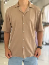 Camisa de manga corta color blanco marfil, confeccionada en tejido texturizado con cuello camisero, cierre frontal de botones y un pequeño bordado oscuro en el pecho.