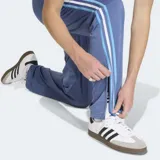 Pantalón de jogging azul con cintura elástica ajustable con cordón, bolsillos laterales con cierre y logo Adidas bordado en dorado. Presenta las tres tiras clásicas de Adidas en blanco y celeste a lo largo de la pierna.