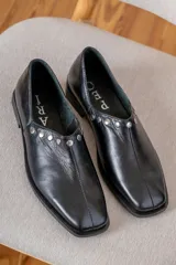 Zapato de cuero vacuno color negro, con punta cuadrada y detalle de tachas metálicas planas en el borde superior. Presenta costura central en el empeine y taco geométrico.