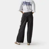 Pantalón de denim negro lavado, corte ancho (loose fit) y tiro alto, con etiqueta rectangular estampada en el muslo derecho y detalle de cinta con colores en el bolsillo delantero izquierdo.