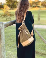 Mochila tipo bucket de gamuza marrón, con detalles de cuero liso en el tono, bolsillo frontal con cierre y cordón ajustable con tiras.