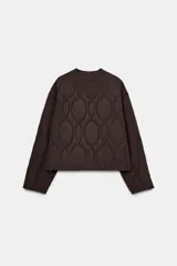 Cazadora corta acolchada de color negro, con cuello redondo, manga larga y cierre frontal con botones. Presenta un diseño de costuras geométricas y bolsillos laterales de vivo.