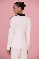 Blazer blanco de sastrería femenina con solapas negras de estilo esmoquin, bordados florales en relieve y detalles de pedrería en hombros y cuerpo.