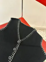 Chaleco tejido color negro con corte sastrero y abotonadura frontal. Presenta un bordado a mano en hilo blanco en todo el contorno de la prenda, incluyendo cuello, sisas y borde inferior.