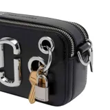 Bandolera Marc Jacobs The Snapshot de cuero negro con herrajes plateados, cierre de doble cremallera, logo metálico frontal y detalles decorativos de anillos, un candado y llaves colgantes.