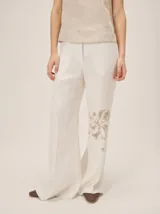 Pantalón de lino color blanco off-white, de corte recto y amplio, con detalle de estampado botánico en tono marrón claro en la parte inferior de una de las piernas.