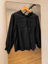 Blusa negra de manga larga con cuello redondo y detalle de volados. Presenta un diseño con frunces en la parte superior y detalles de encaje horizontal en el pecho.